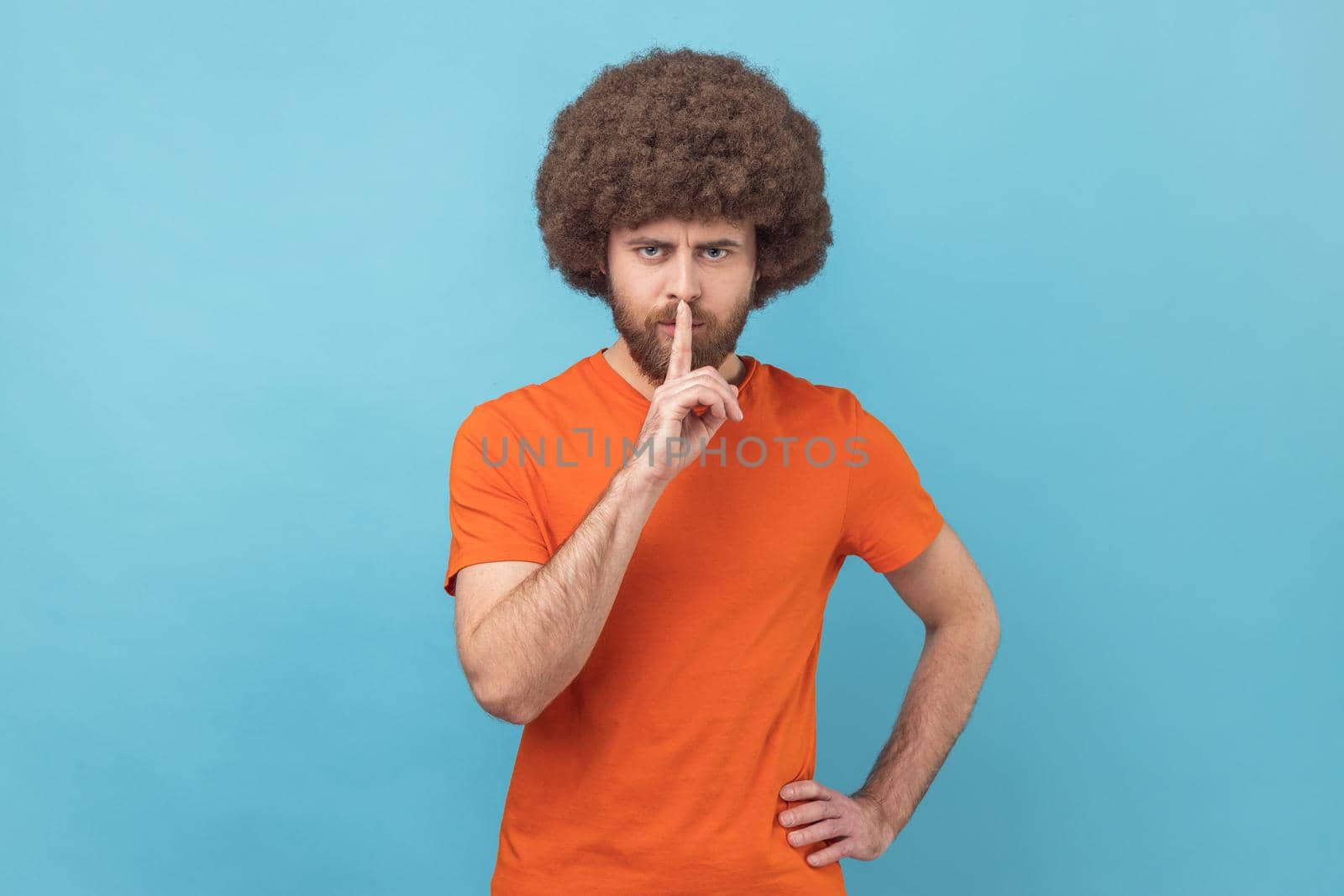 Portrait of man with Afro hairstyle wearing orange T-shirt holding finger on lips making hush silence gesture, asking to keep secret, don't speak. Indoor studio shot isolated on blue background.