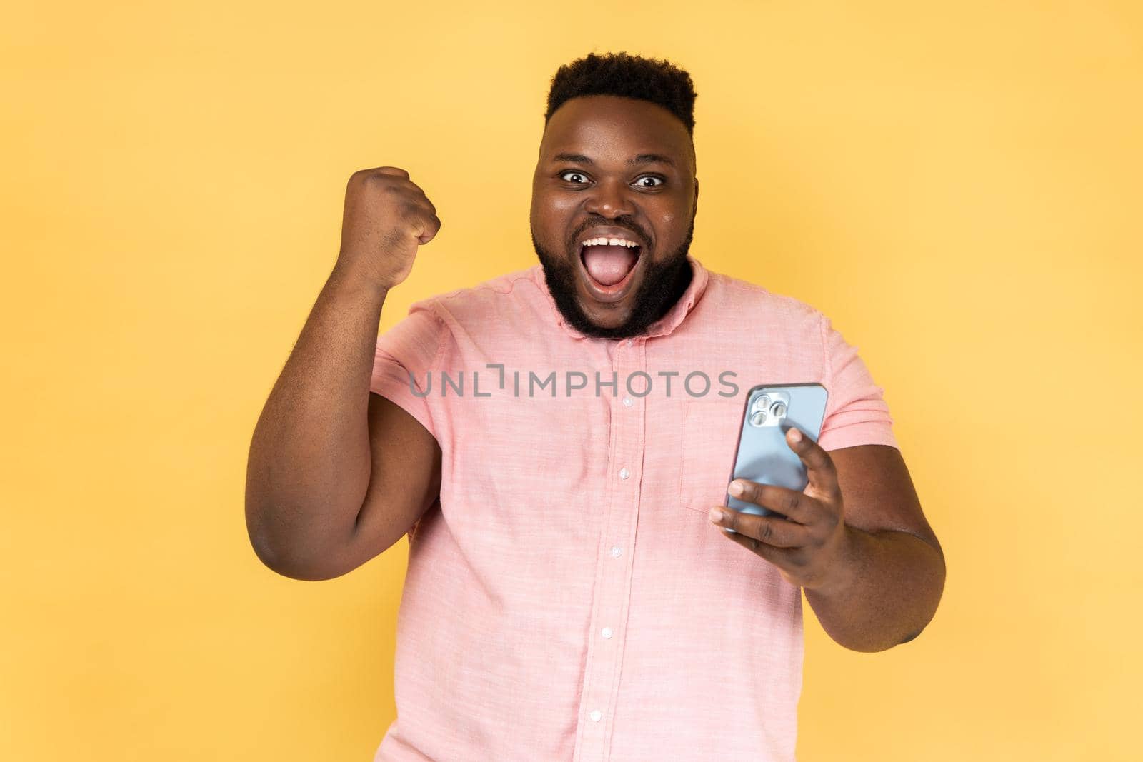 Portrait of satisfied bearded man wearing pink shirt standing with cell phone in hands and clenching fist, celebrating his victory, looks at camera. Indoor studio shot isolated on yellow background.