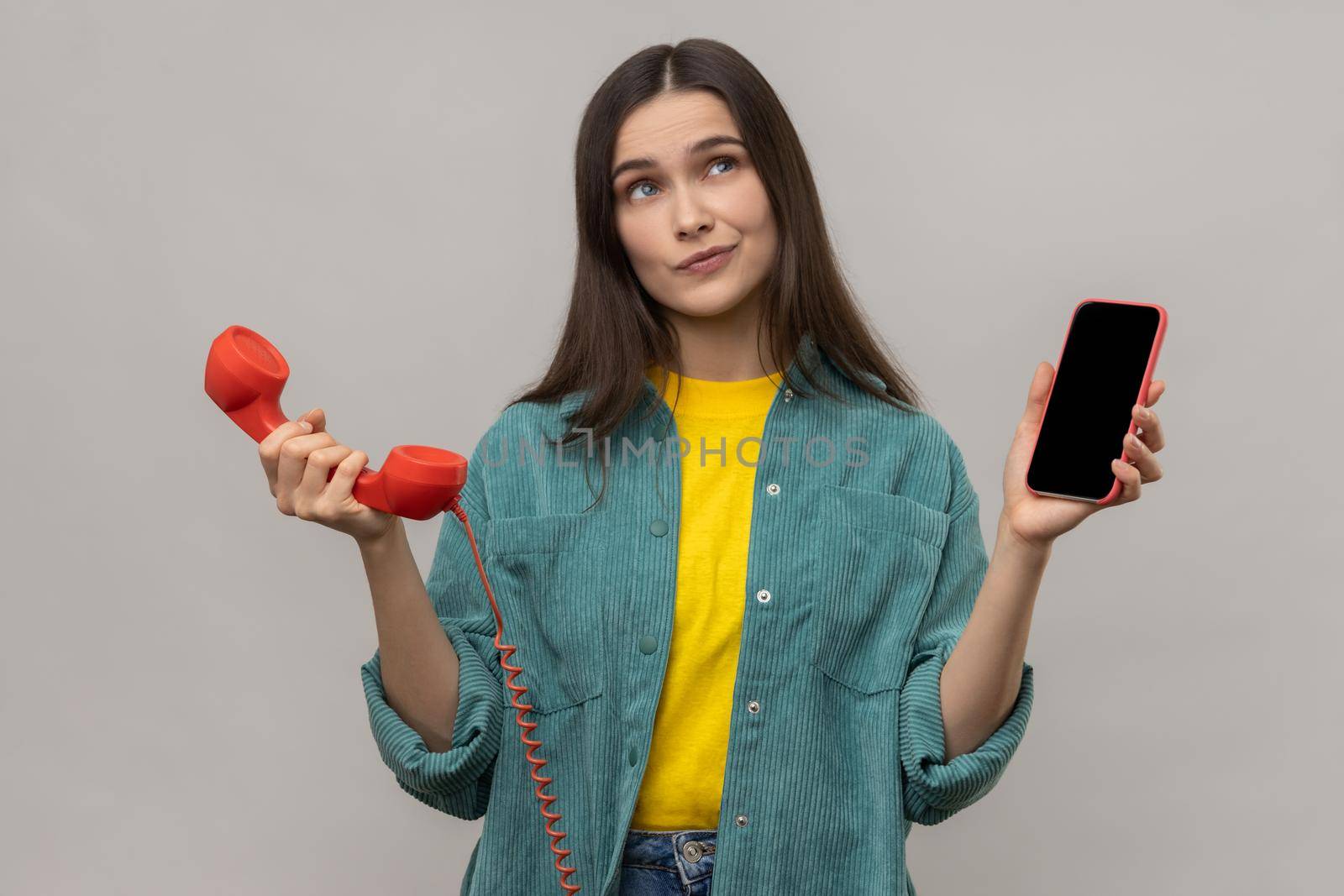 Portrait of confused puzzled woman standing retro telephone and mobile phone, thinking what to use better, wearing casual style jacket. Indoor studio shot isolated on gray background.