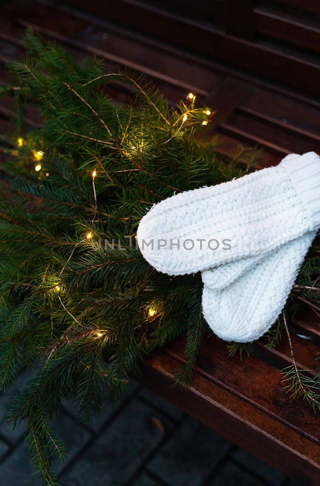 Branches of a coniferous tree decorated with a garland lie on a bench along with knitted white mittens. Place for an inscription