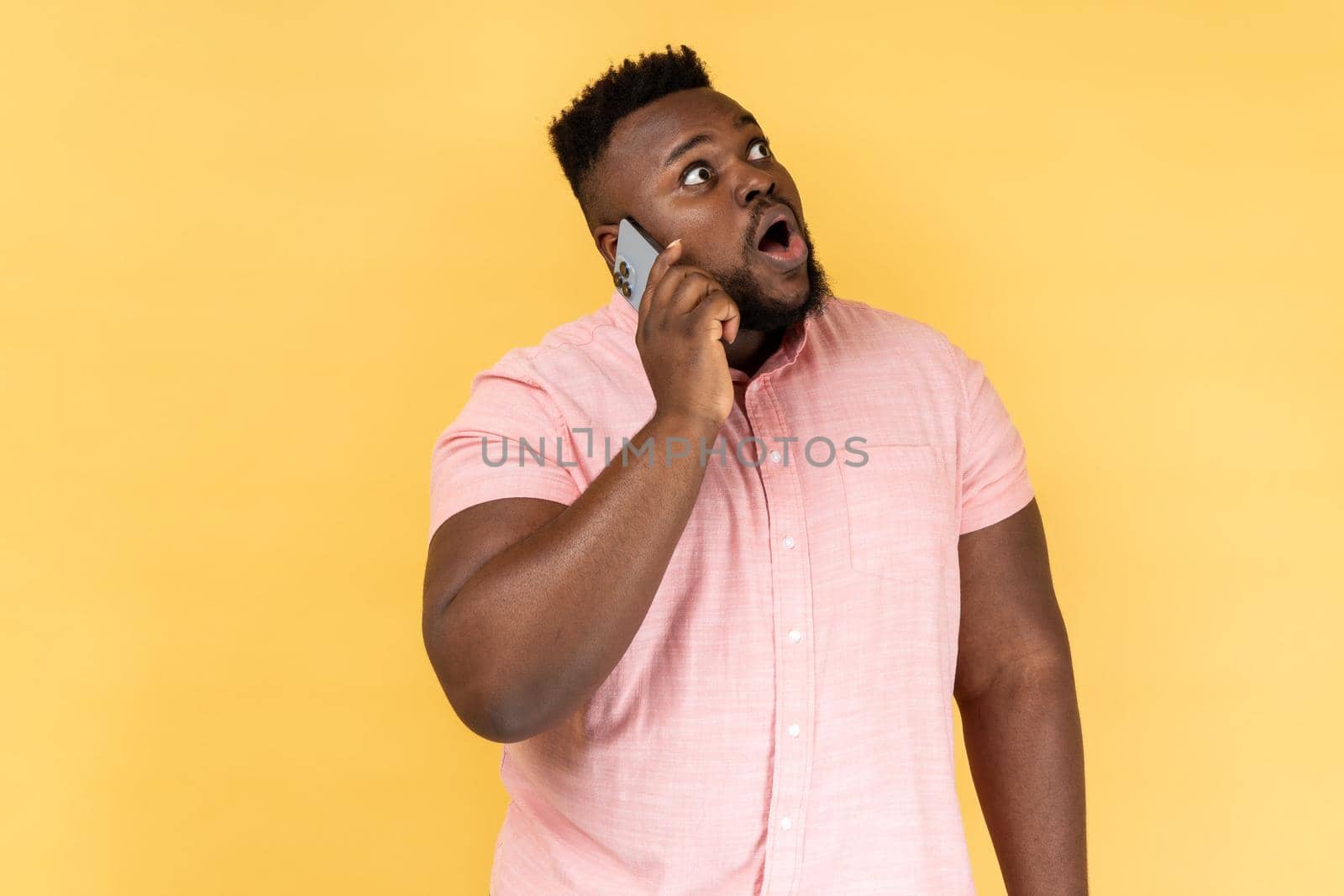 Portrait of shocked man wearing pink shirt talking with friend on cell phone, looking away with surprised amazed, hearing breaking news. Indoor studio shot isolated on yellow background.