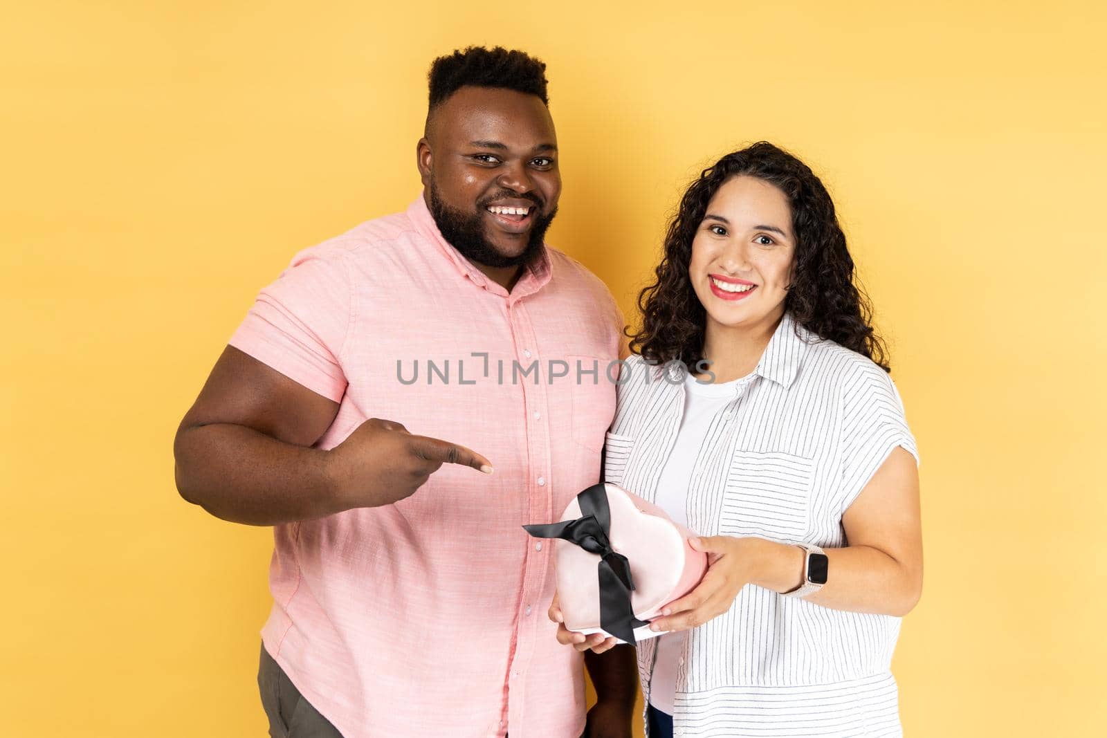 Portrait of delighted smiling young couple in casual clothing standing together, man pointing at present box in her wife hands, celebrating holiday. Indoor studio shot isolated on yellow background.
