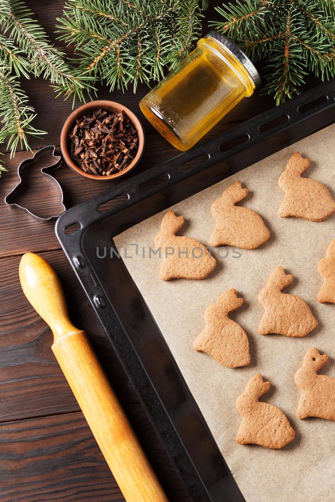 Ready-made Christmas cookies in the form of a rabbit on a baking sheet on a wooden table decorated with Christmas tree branches, with a jar of honey and cloves. Symbol of 2023.Vertical photo