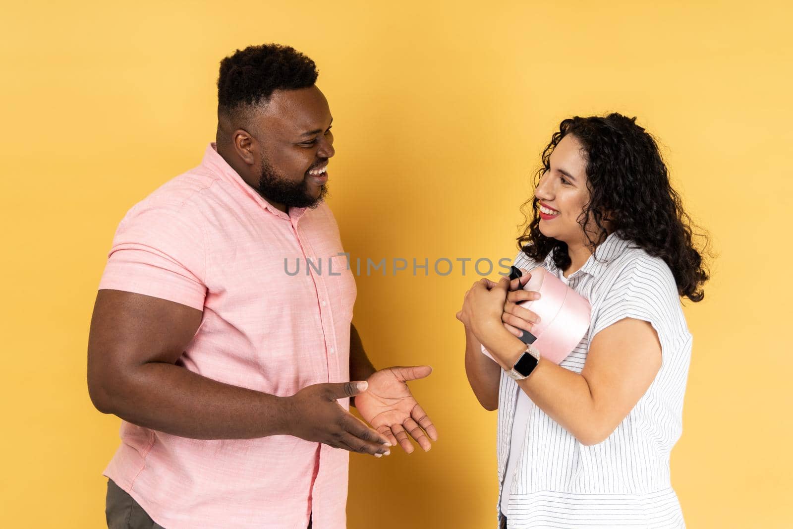 Portrait of happy young couple in casual clothing standing together, woman holding present box, a gift from her husband, celebrating anniversary. Indoor studio shot isolated on yellow background.