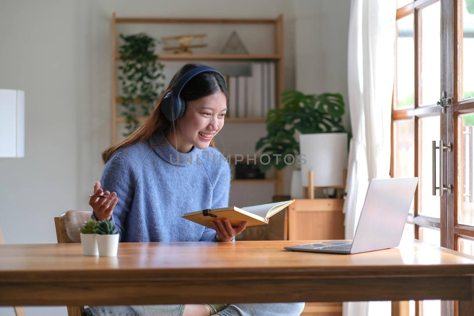 Smiling young asian teen girl wear headphones video calling on laptop. Happy pretty woman student looking at computer screen watching webinar or doing video chat by webcam. by wichayada