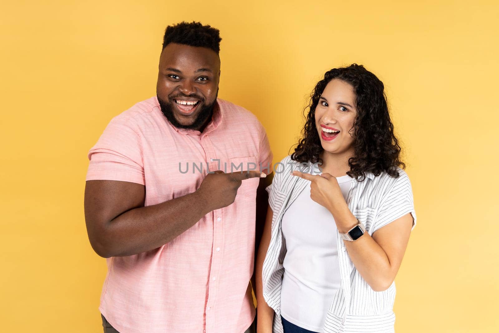 Portrait of happy positive young couple in casual clothing standing together, pointing fingers at each other and looking smiling at camera. Indoor studio shot isolated on yellow background.