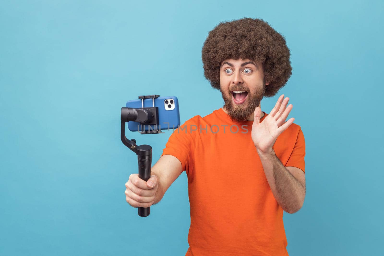 Portrait of man with Afro hairstyle wearing orange T-shirt using stedicam and phone for livestream, waving hand to followers, content for business vlog. Indoor studio shot isolated on blue background.
