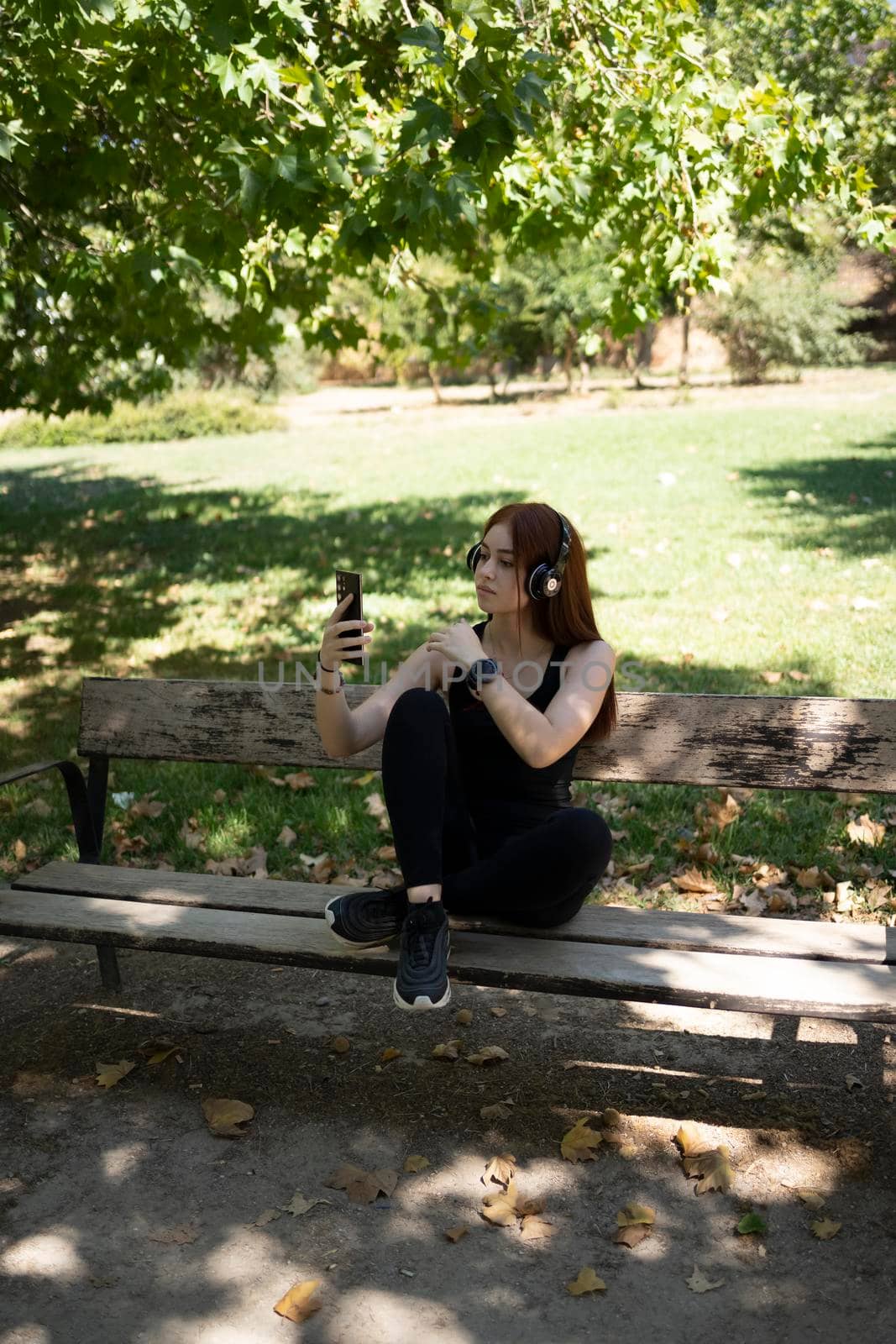Full body young female athlete browsing cellphone and listening to music in wireless headphones while taking break in running session on sunny summer day in park