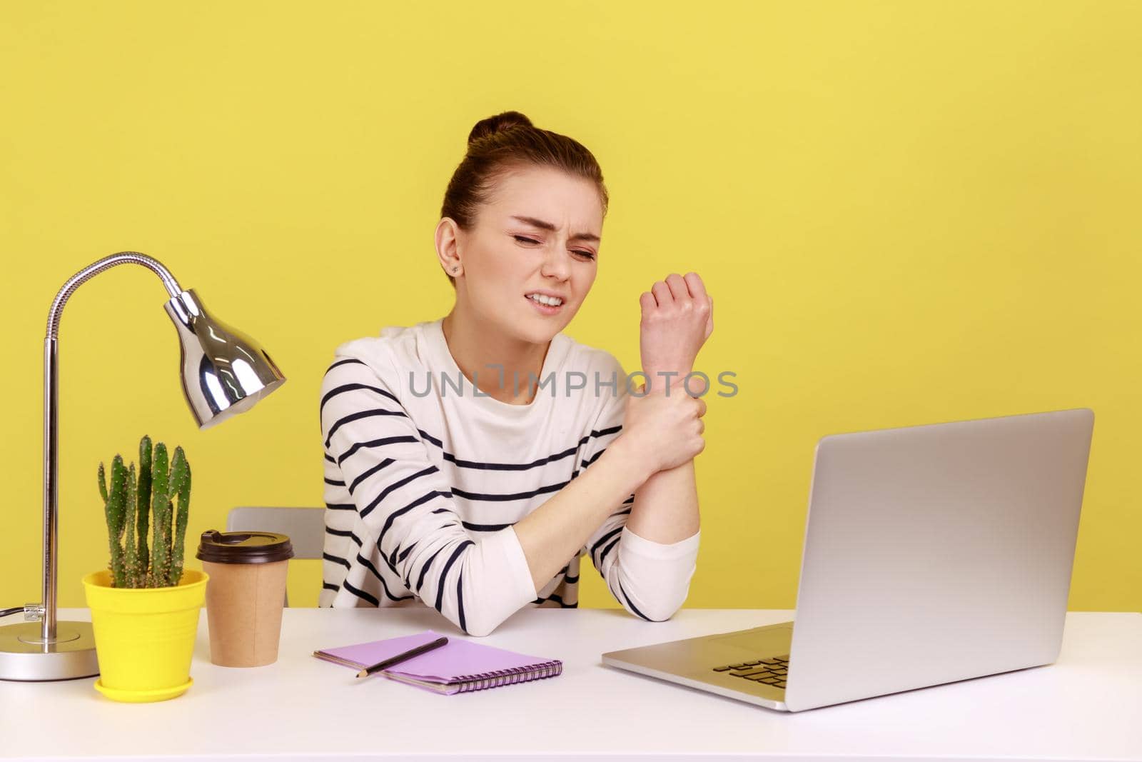 Tired sick woman office manager sitting at workplace with laptop and holding sore wrist, suffering pain in injured hand, carpal tunnel syndrome. Indoor studio studio shot isolated on yellow background
