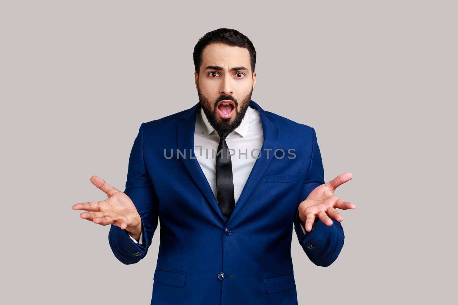 Portrait of bearded man standing with raised hands and indignant expression, asking reason of conflict, wearing official style suit. Indoor studio shot isolated on gray background.