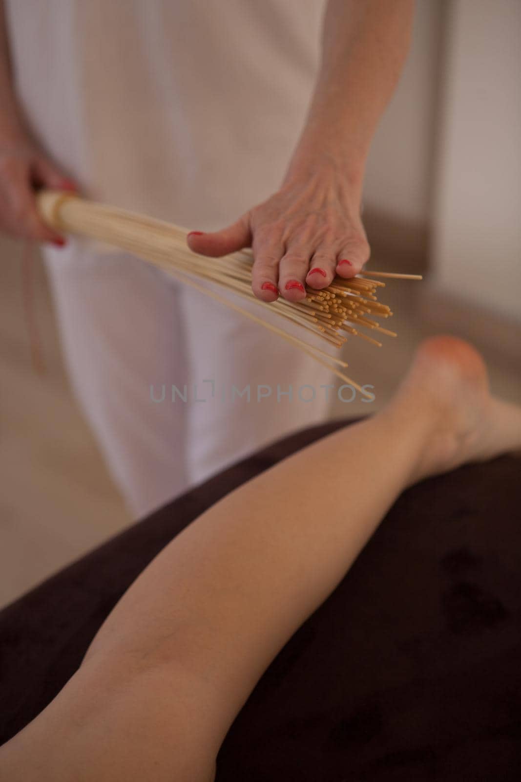 Vertical shot of masseur using bamboo massage broom on legs of female client