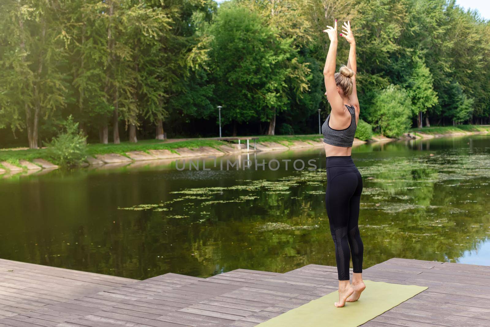 A slender woman stands on a green rug, on a wooden platform by a pond in the park, in summer, with her hands up, doing yoga. Copy space