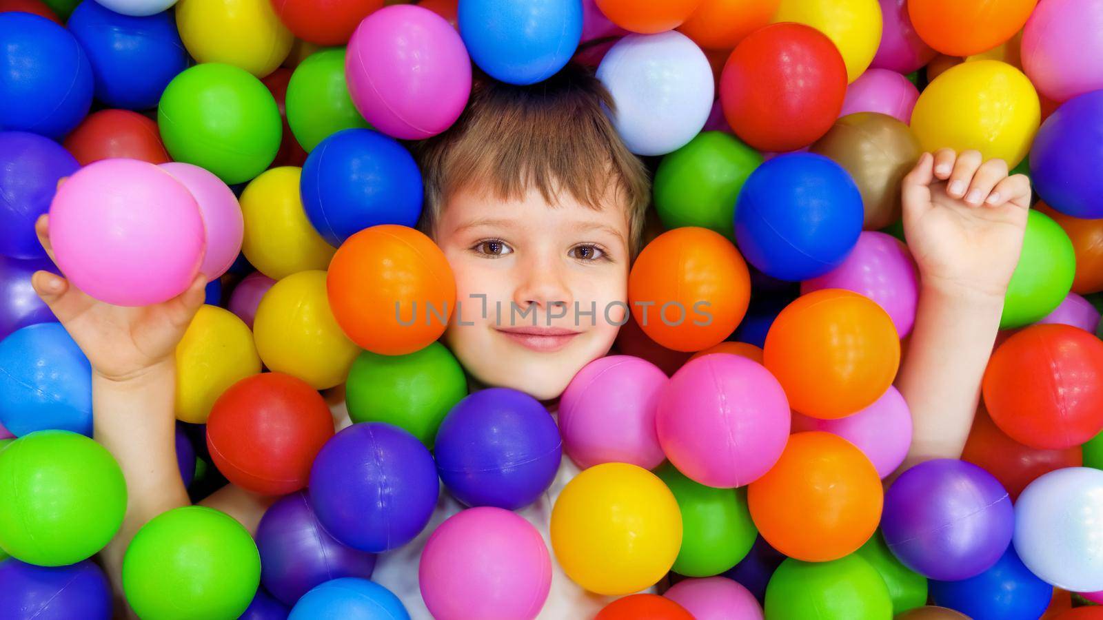 Smile kid lying colorful plastic balls pool. Colorful balls dry pool kindergarten playground child indoor play area. Playroom kids ball pit. Caucasian boy indoor playground kids play zone or kids zone by synel