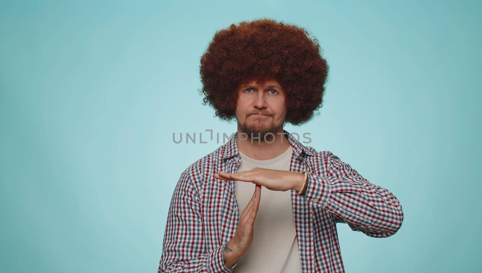 Tired serious upset man 30s with lush hairstyle wig showing time out gesture, limit, stop break sign, no pressure, I need more time. Young adult guy boy isolated alone on blue studio wall background