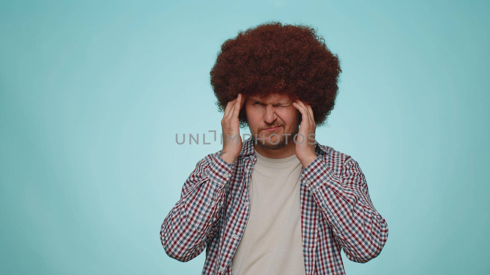 Bearded young adult man in shirt rubbing temples to cure headache problem, suffering from tension and migaine, stress, grimacing in pain, high blood pressure isolated alone on blue wall background