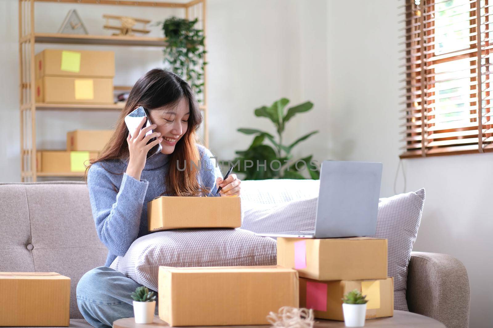Portrait young asian woman using laptop computer for work with parcel box on sofa in living room interior.