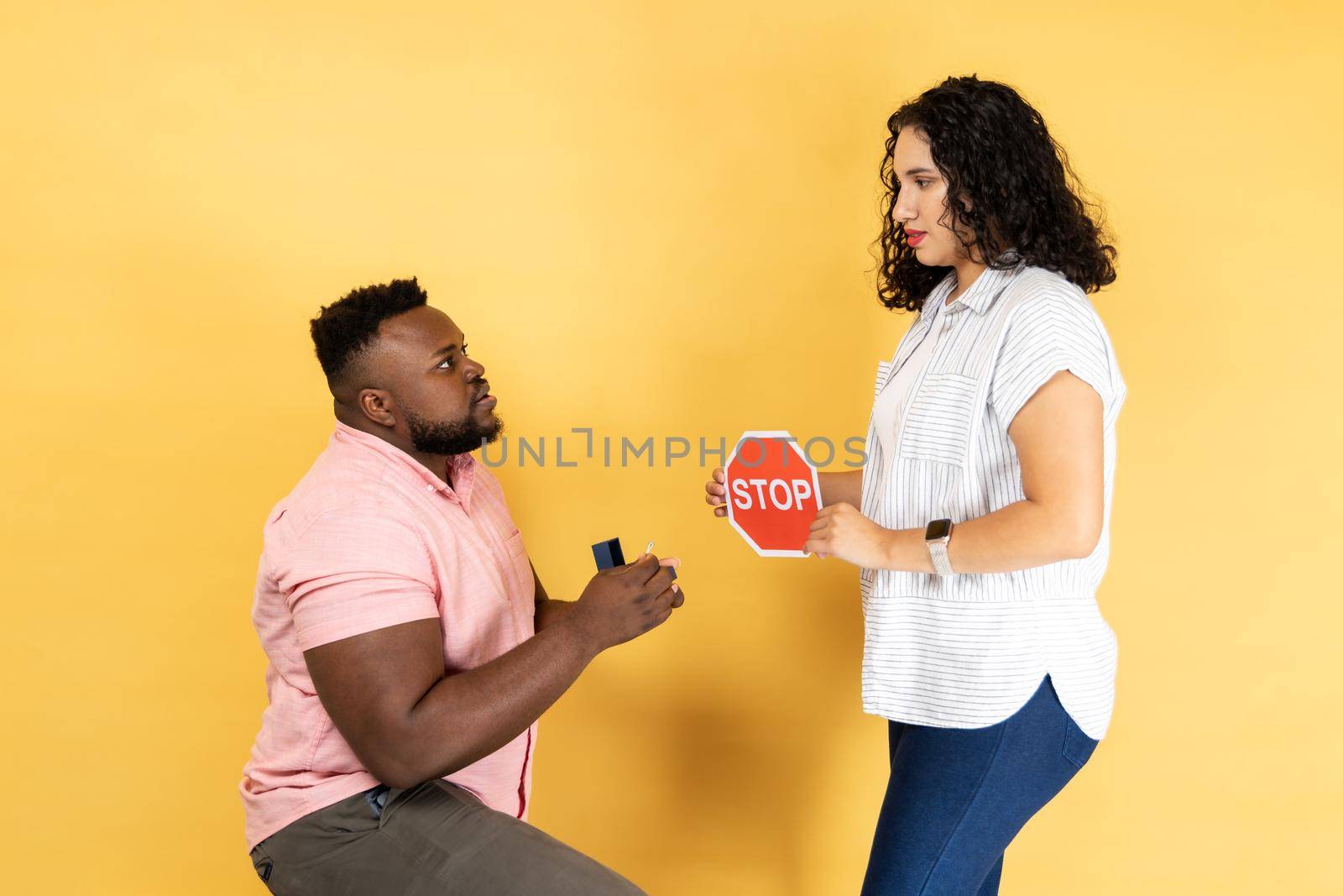 Portrait of young couple in casual clothing standing together, man making proposal to his girlfriend, woman showing red stop sign to him, refusing. Indoor studio shot isolated on yellow background.