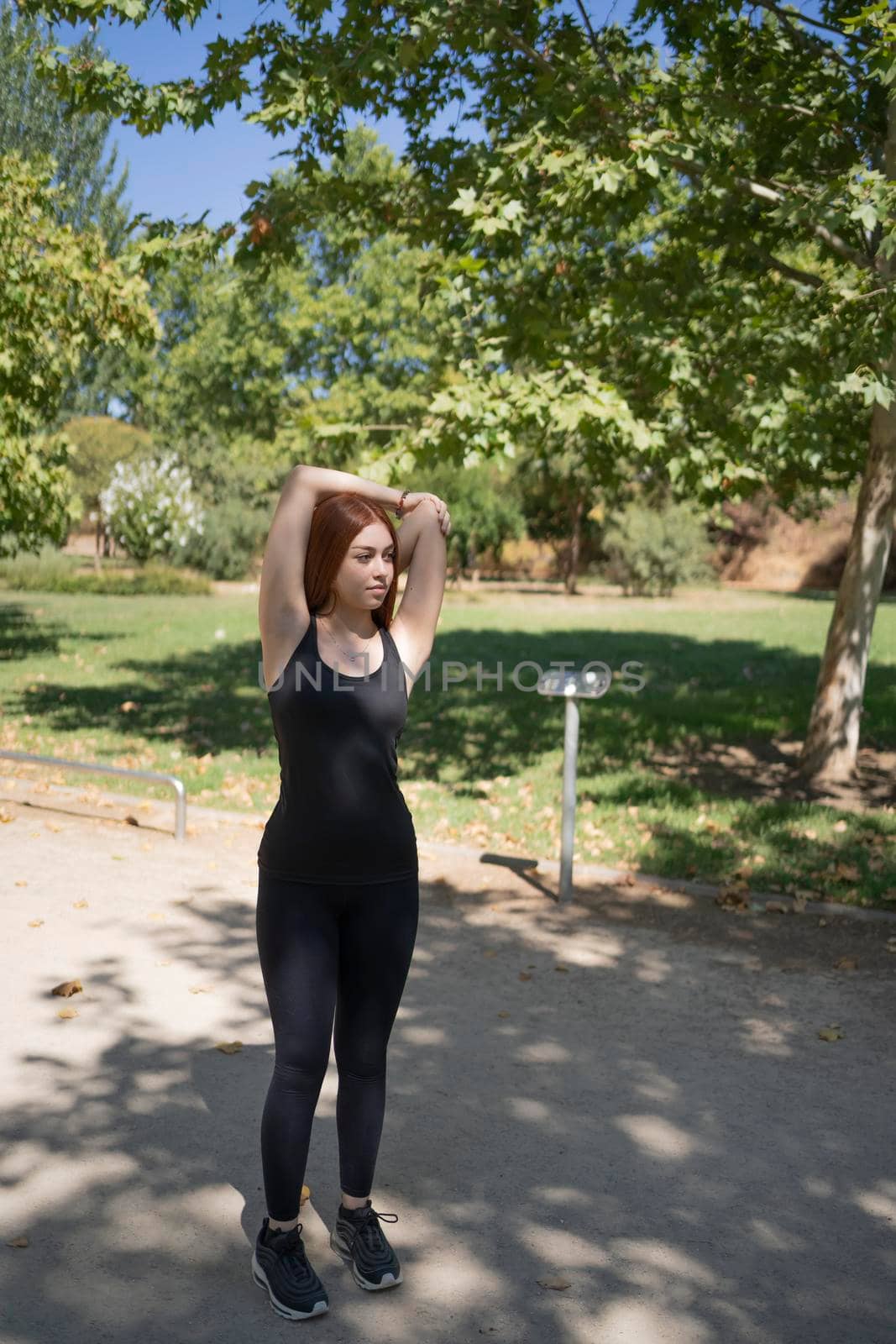Fitness young redhead woman doing stretching exercise outdoors in public park. by barcielaphoto