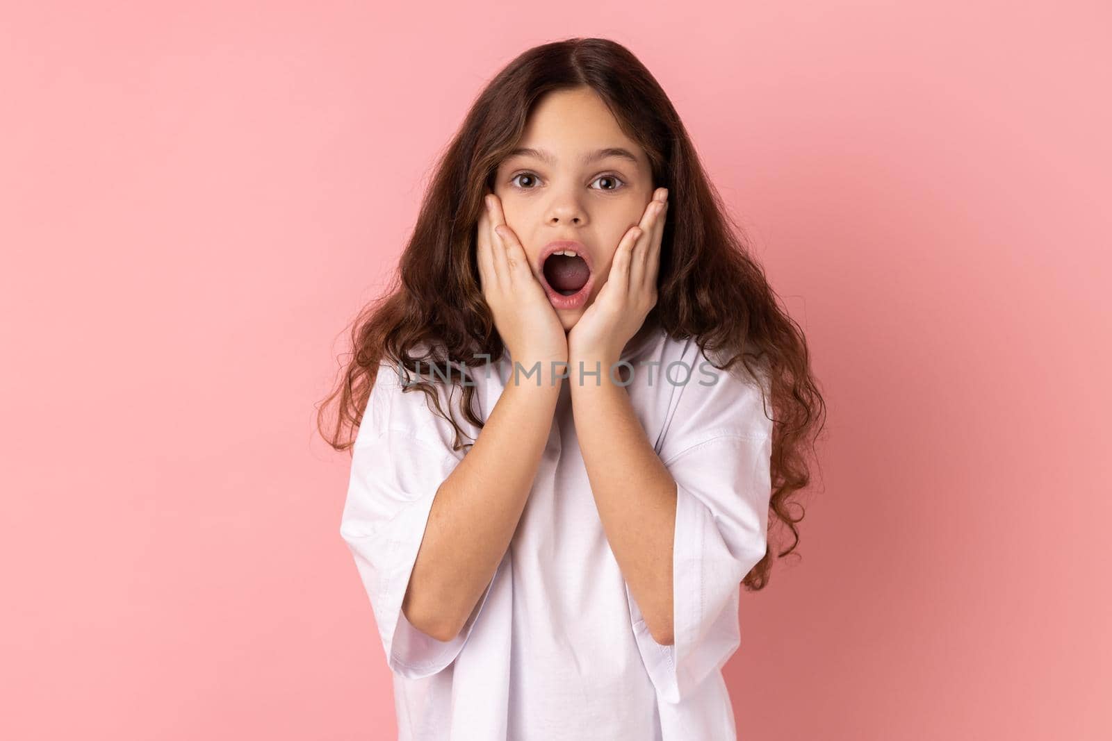 Portrait of amazed little girl wearing white T-shirt standing with hands on cheeks, looking at camera with big eye, saying wow. Indoor studio shot isolated on pink background.