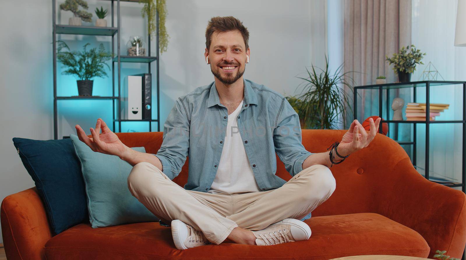 Keep calm down, relax, inner balance. Adult man breathes deeply with mudra gesture, eyes closed meditating with concentrated thoughts, peaceful mind. Young guy sit at home in living room on couch