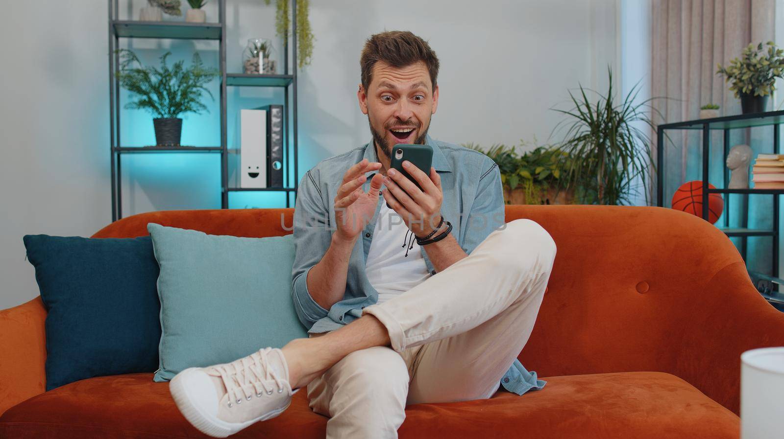 Oh my God Wow. Excited happy joyful winner man use smartphone typing browsing found out great win good news celebrate victory. Young guy with mobile phone sitting at home in room on orange couch