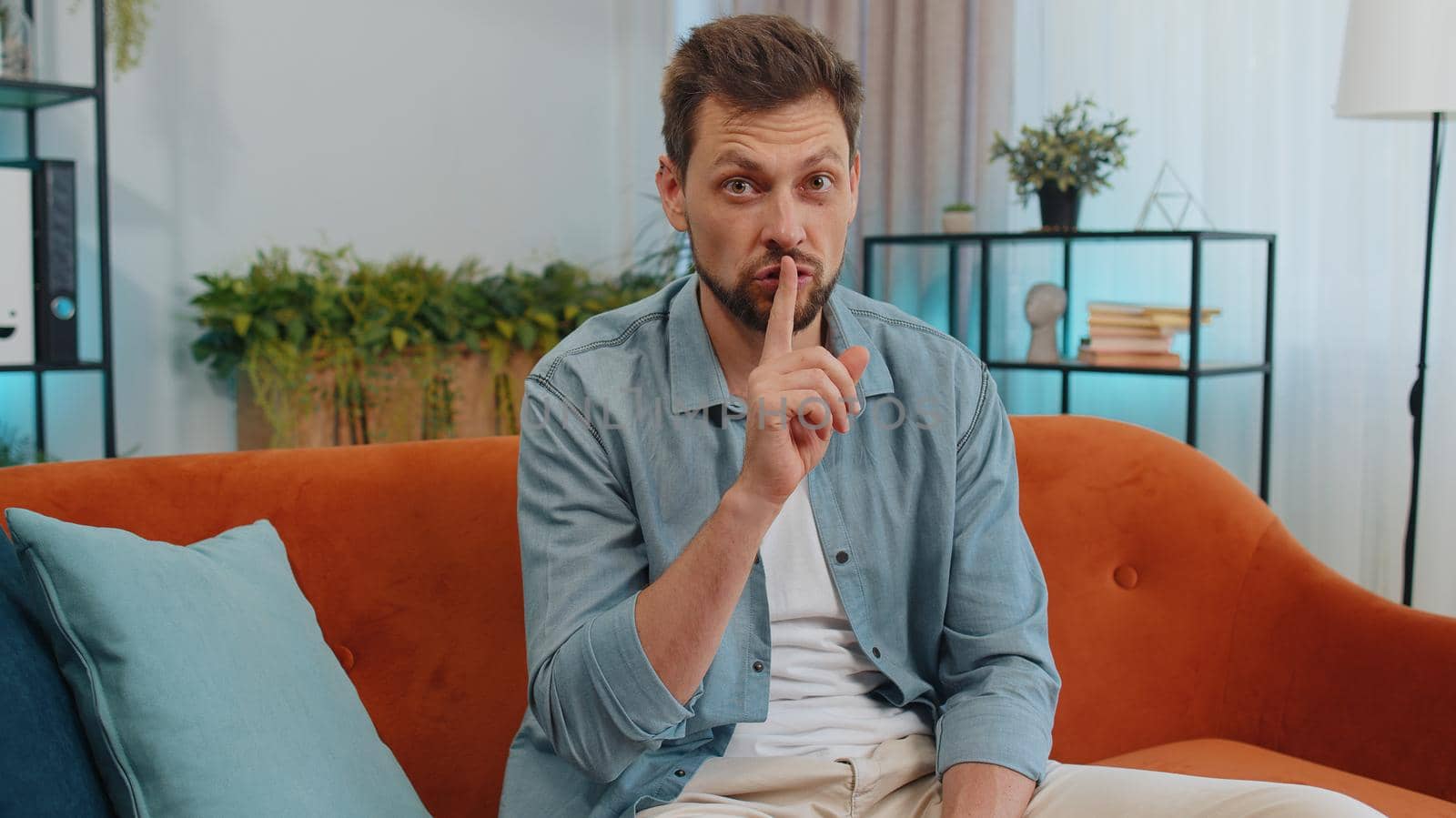 Shh be quiet please. Adult caucasian man presses index finger to lips makes silence hush gesture sign do not tells gossip secret. Young guy at home apartment living room sitting on orange couch