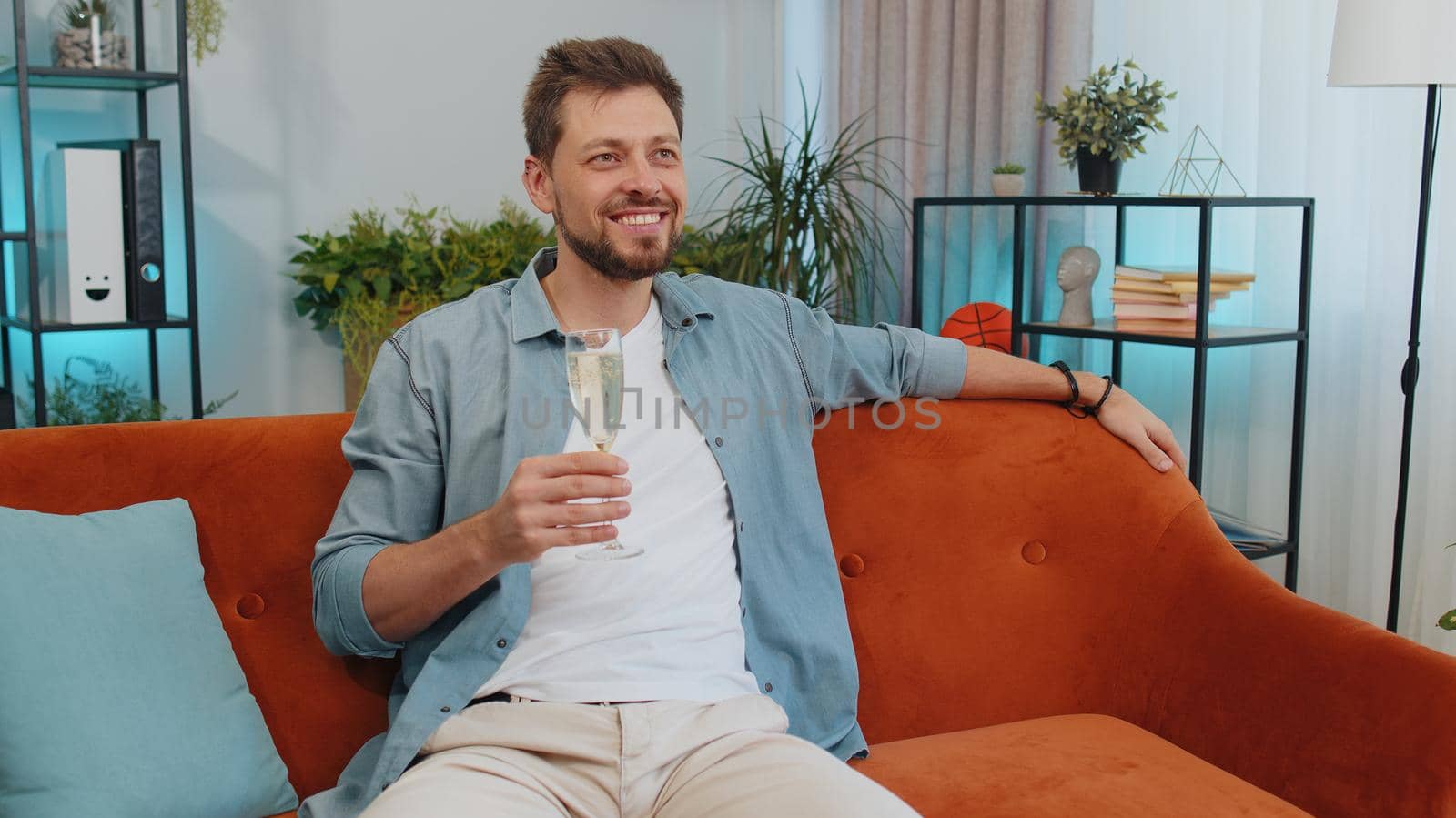 Joyful caucasian adult man hold glass of champagne cheering drinking, celebrate success win, career grow up, lucky situation at home in living room. Young guy celebrating rent or purchase of new house