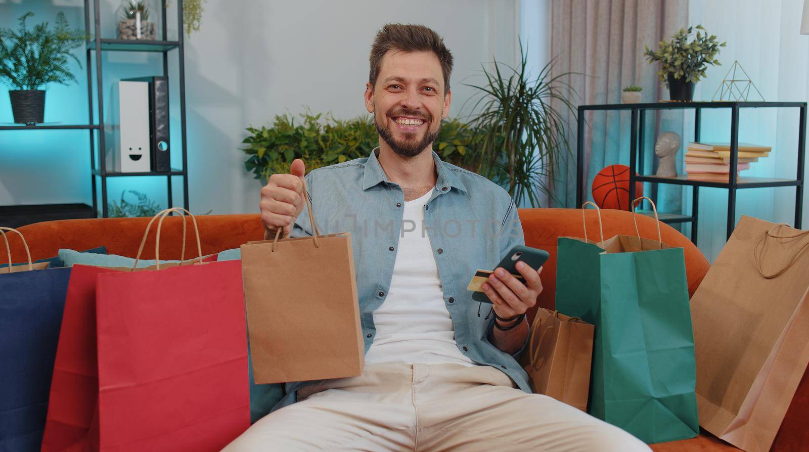 Happy shopaholic consumer man sitting with shopping bags at modern home apartment making online order, payment with credit plastic bank card. Young guy on couch at home. Discount sale on internet