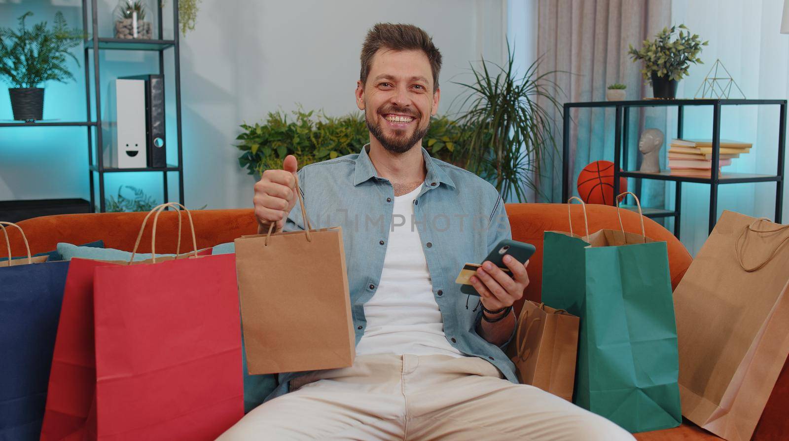 Happy shopaholic young man sitting with shopping bags making online payment with credit card by efuror