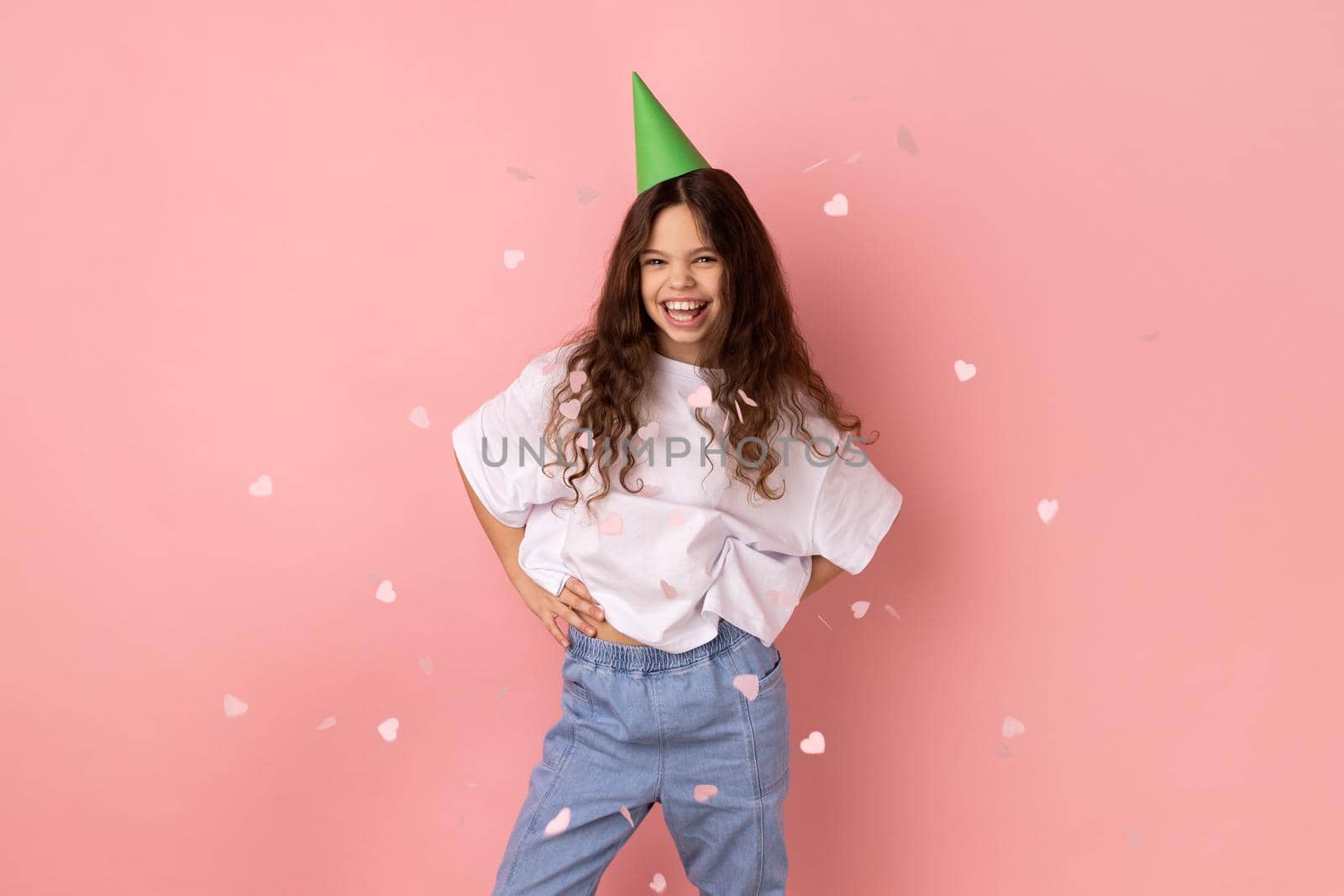 Little girl wearing white T-shirt and party cone standing under falling confetti, looking at camera smiling, celebrating birthday holiday, festive mood. Indoor studio shot isolated on pink background.
