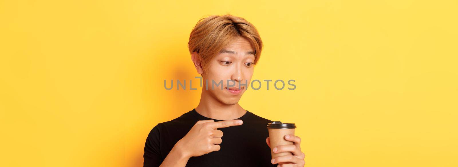 Close-up of impressed handsome asian guy pointing finger at delicious coffee, standing amazed over yellow background by Benzoix
