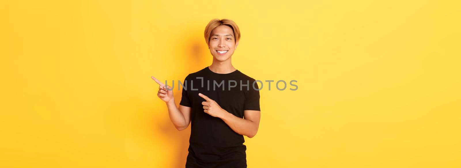 Portrait of satisfied smiling blond guy, asian student pointing fingers right, showing way, standing yellow background by Benzoix