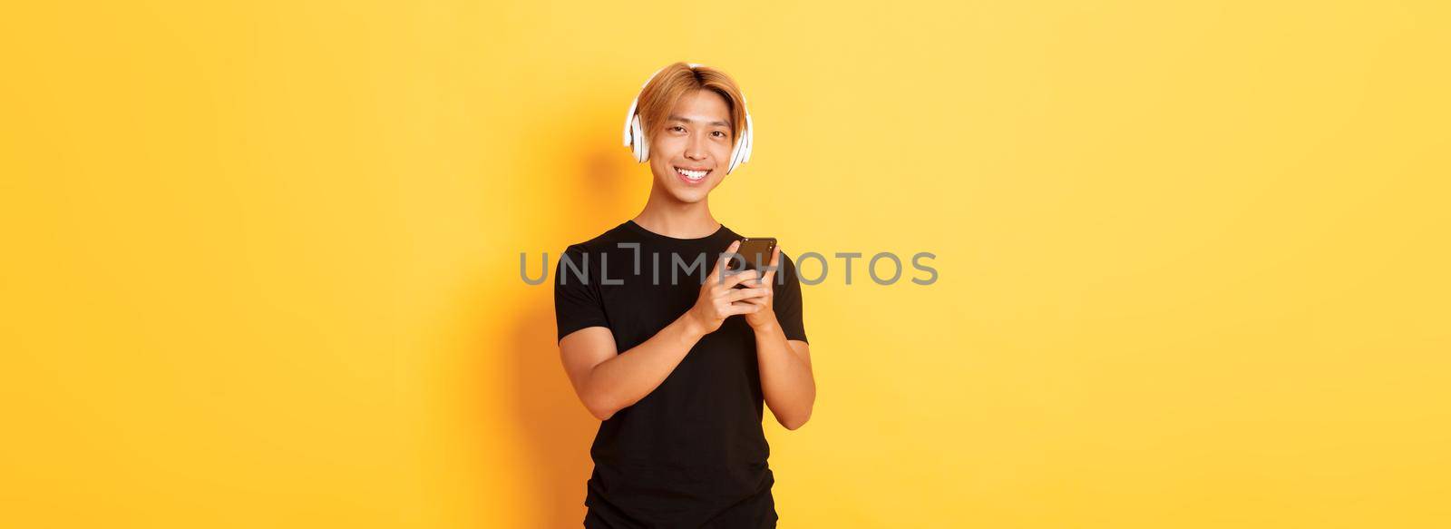Good-looking smiling, pleased asian guy in wireless headphones, holding smartphone and looking satisfied camera, listening music or podcast.