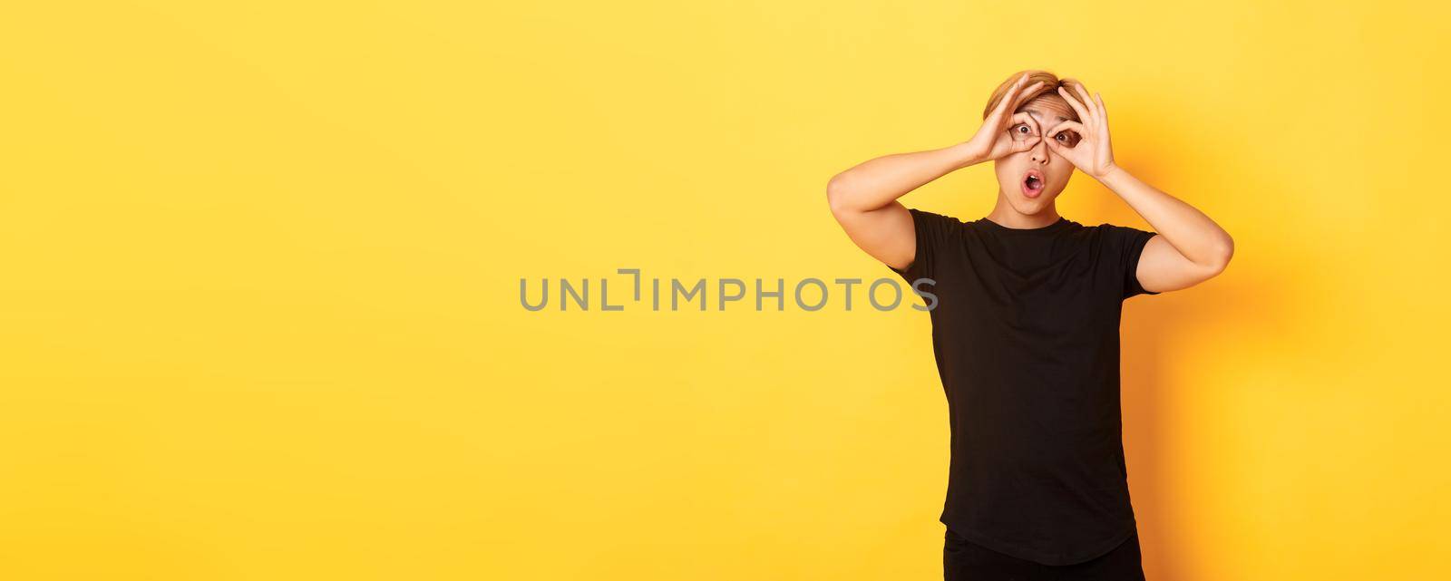 Portrait of funny and cute asian blond guy making fingers glasses and grimacing, standing yellow background.