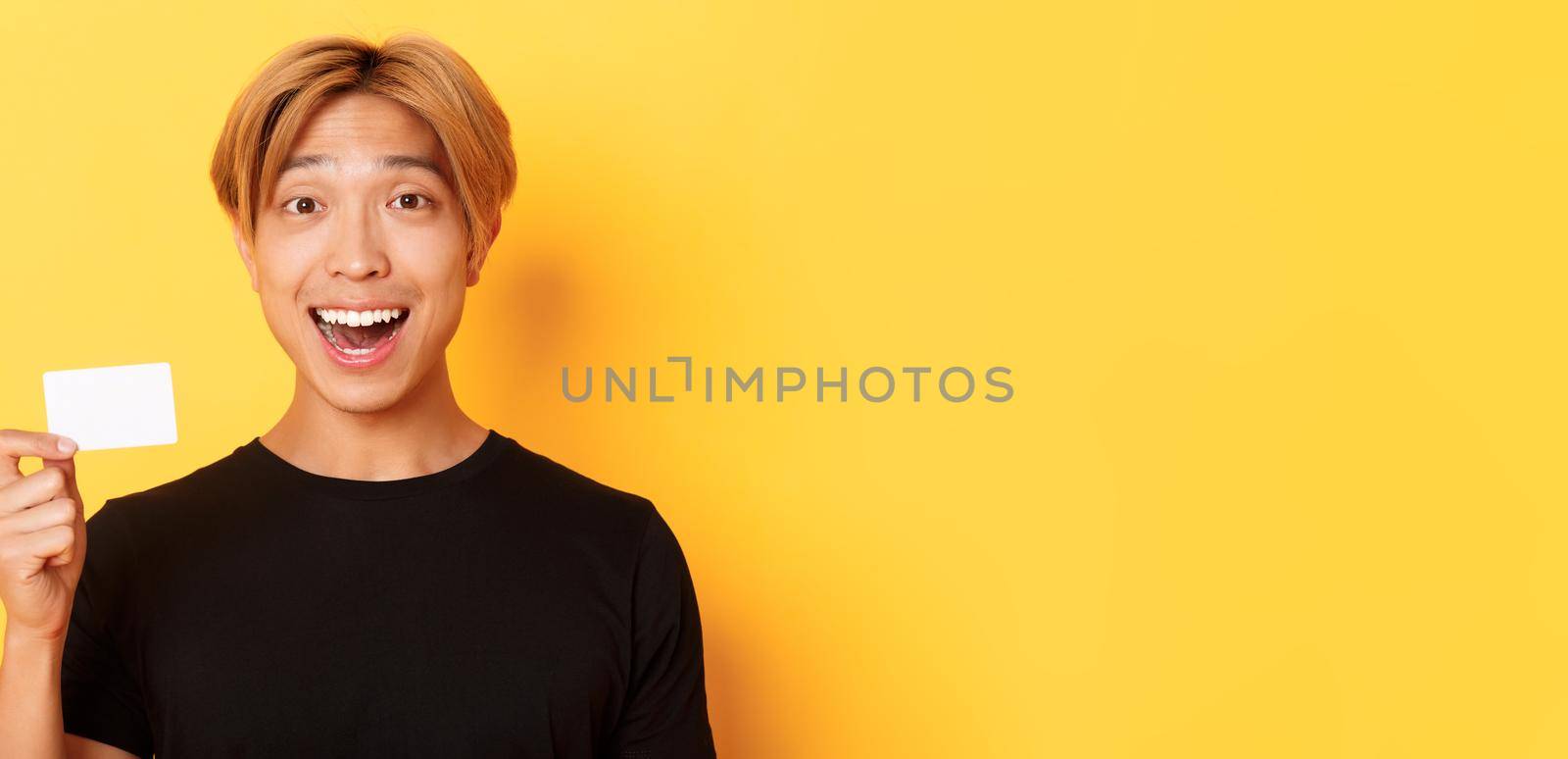Close-up of excited surprised handsome asian guy, smiling amazed and showing credit card, standing yellow background.