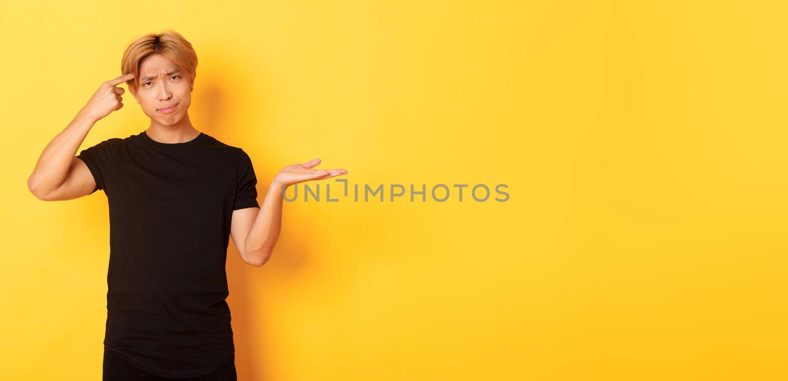 Disappointed asian guy with blond hair, raising hand up puzzled and scolding someone acting stupid, standing yellow background.
