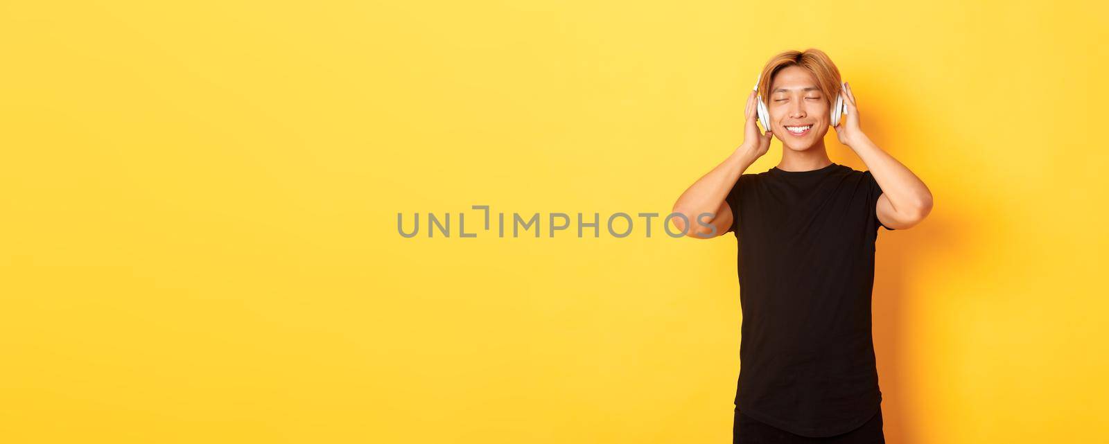 Relaxed and pleased handsome asian guy close eyes from satisfaction while listening music in wireless headphones, standing yellow background by Benzoix