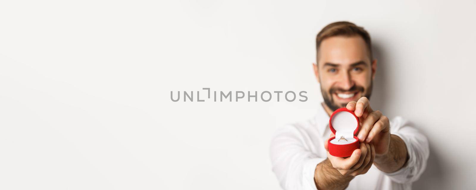 Close-up of handsome man asking to marry him, focus on box with wedding ring, concept of proposal and relationship, white background.