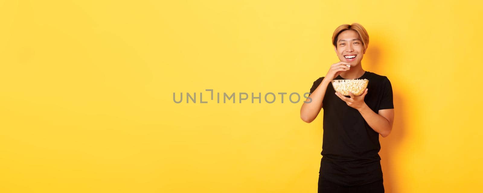 Carefree happy asian guy with blond hair, watching comedy, laughing and eating popcorn, standing yellow background by Benzoix