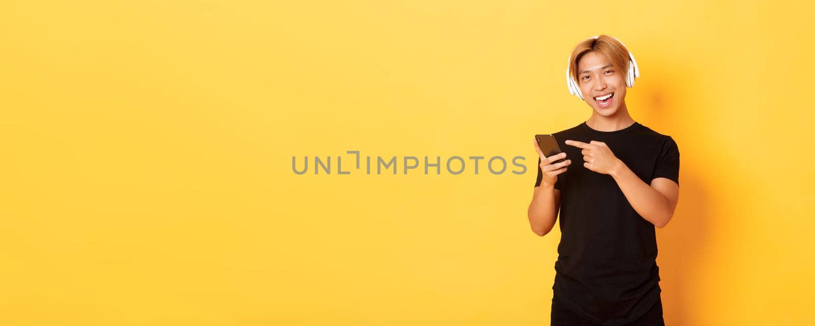 Joyful satisfied asian handsome guy, listening music or good podcast in headphones, pointing finger at smartphone with pleased smile, yellow background.