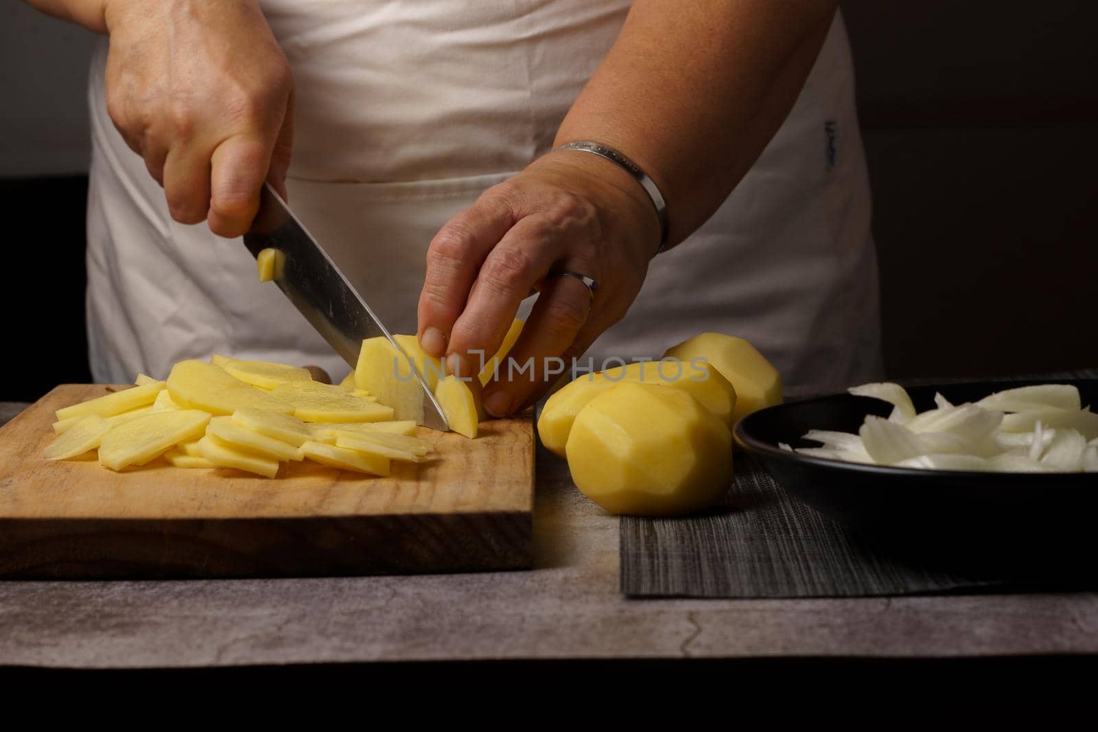 woman slicing potatoes with a knife on a wooden board next to a black plate with sliced onion