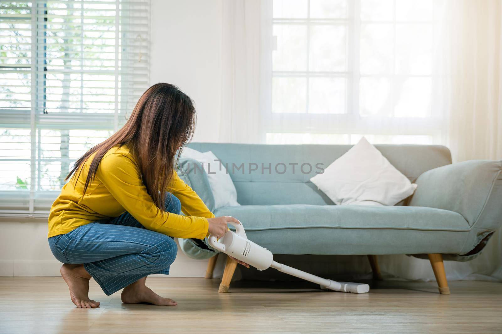 Housewife female dust cleaning floor under sofa or couch furniture with vacuum cleaner, Happy Asian young woman with accumulator vacuum cleaner at home in living room, household and housework concept