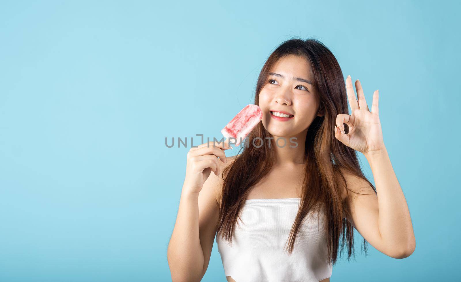 Sweet tasty frozen dessert on summer time. Happy Asian young beautiful woman holding delicious ice cream wood stick mixed fruit flavor and show OK sign, studio shot isolated on blue background