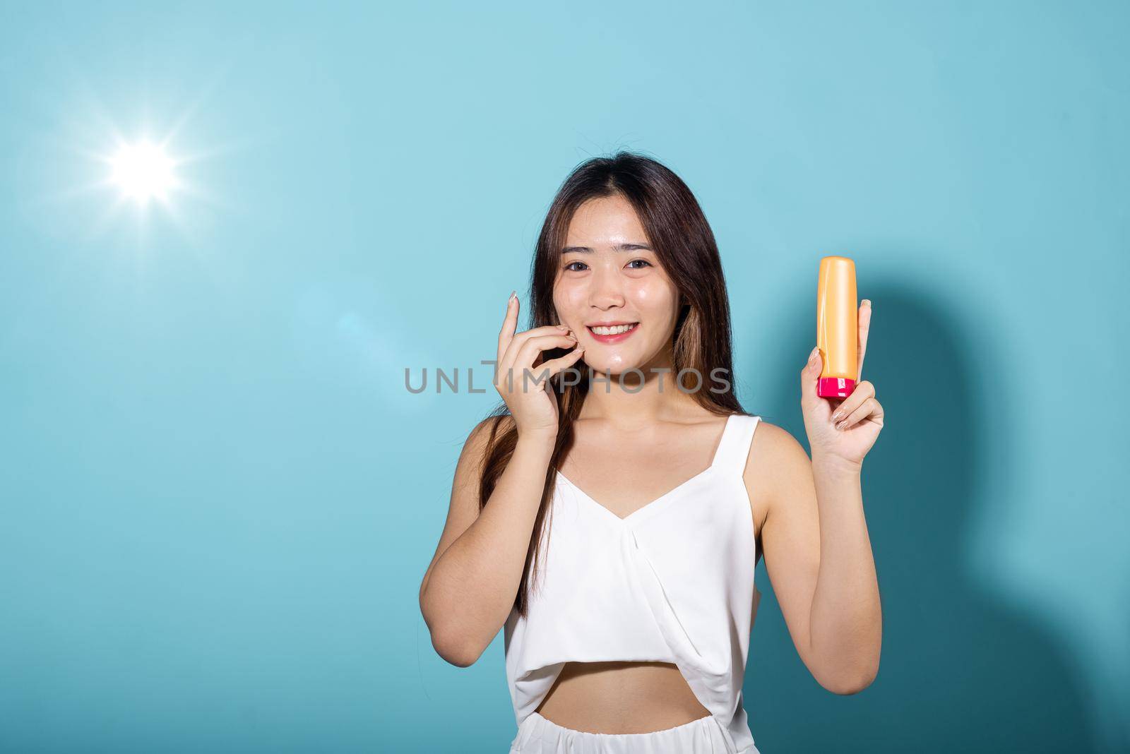 Beautiful brunette female hold cream sunblock bottle in studio shot isolated on blue background, Asian young woman holding body lotion on hand, tube of sunscreen, Beauty cosmetics facial treatment
