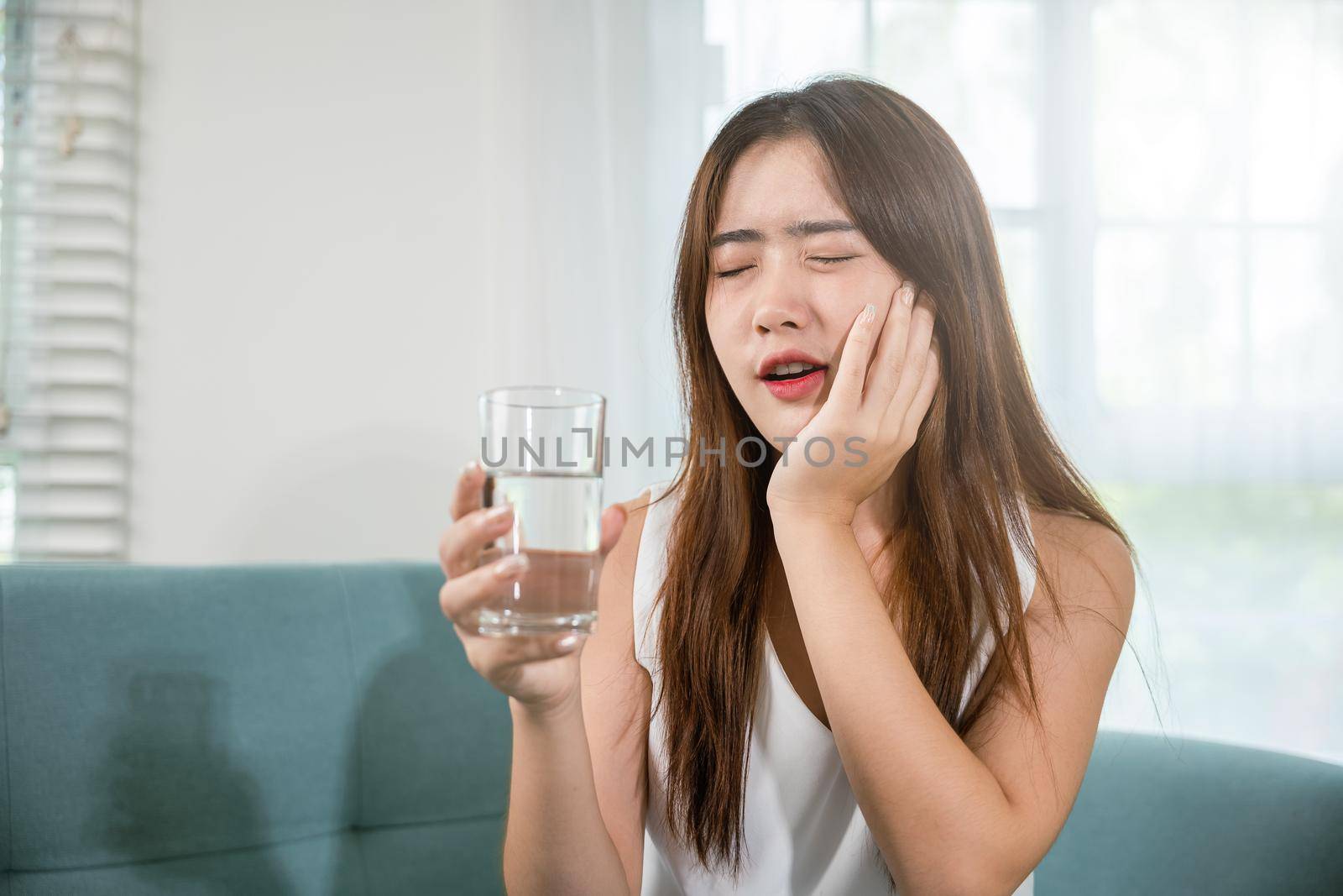 Beautiful female toothache and dental problems touching cheek feeling pain after drinking cold water, Asian young woman with sensitive teeth holding glass of cold water at home in living room, pain