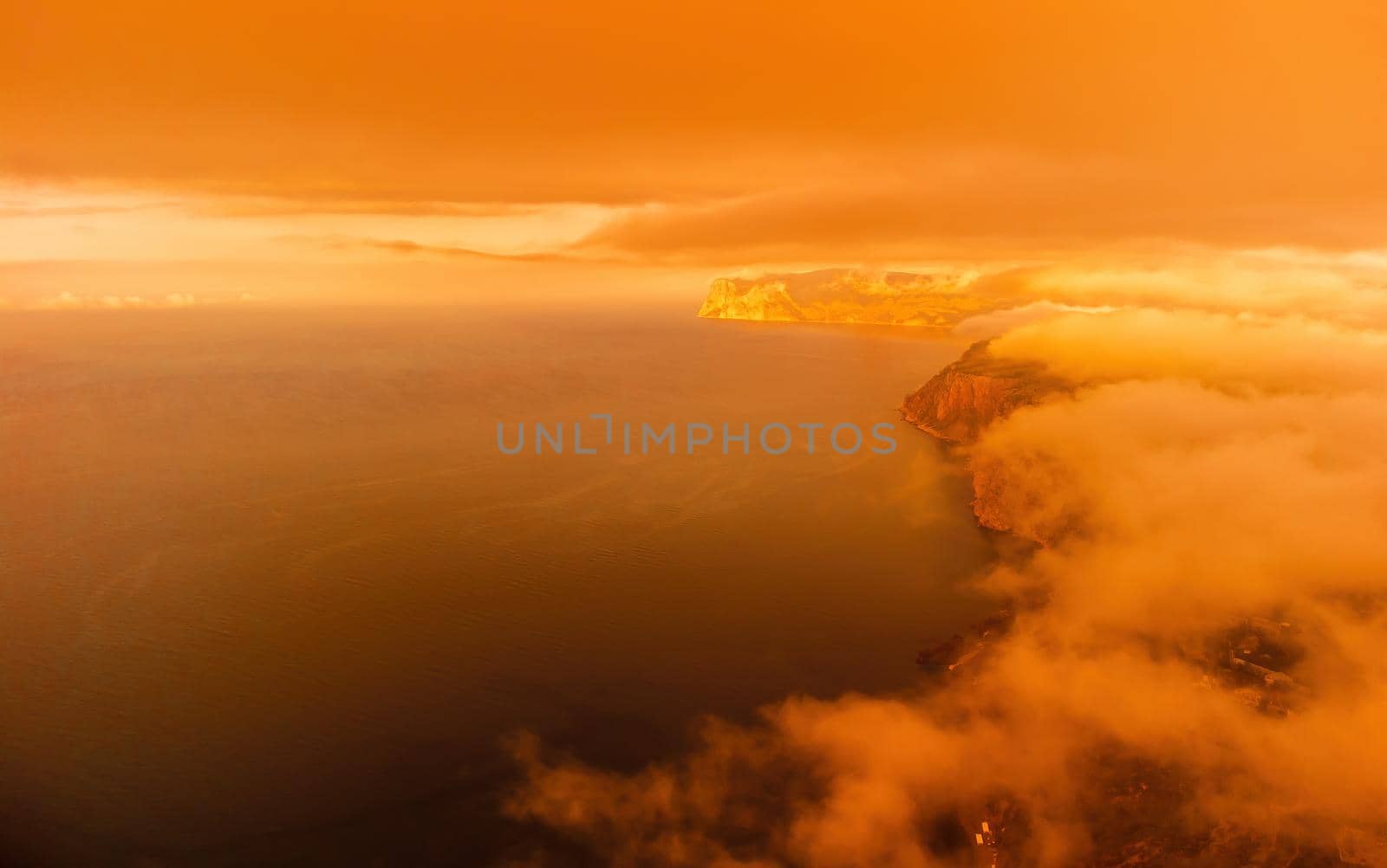 A red burning sunset with the silhouette of a cliff over the sea. Abstract nature summer or spring ocean sea background. Small waves on golden warm water surface with bokeh lights from sun.