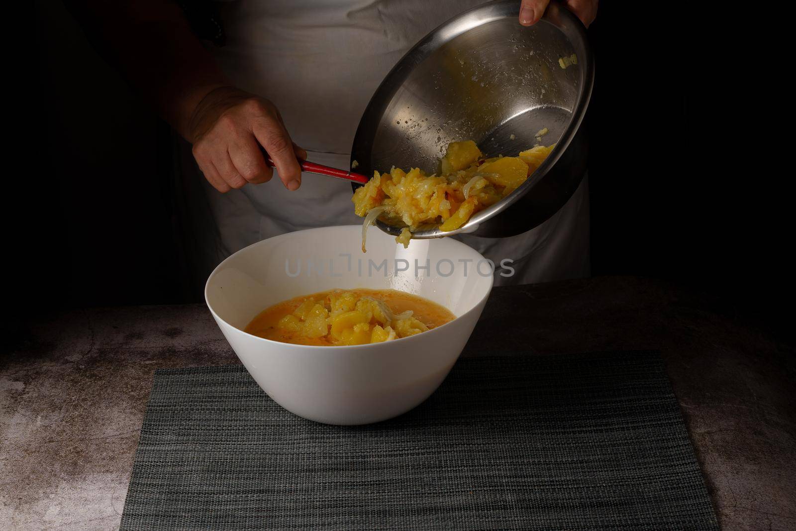 woman in white apron mixing the ingredients of a potato omelet in a white bowl, dark background