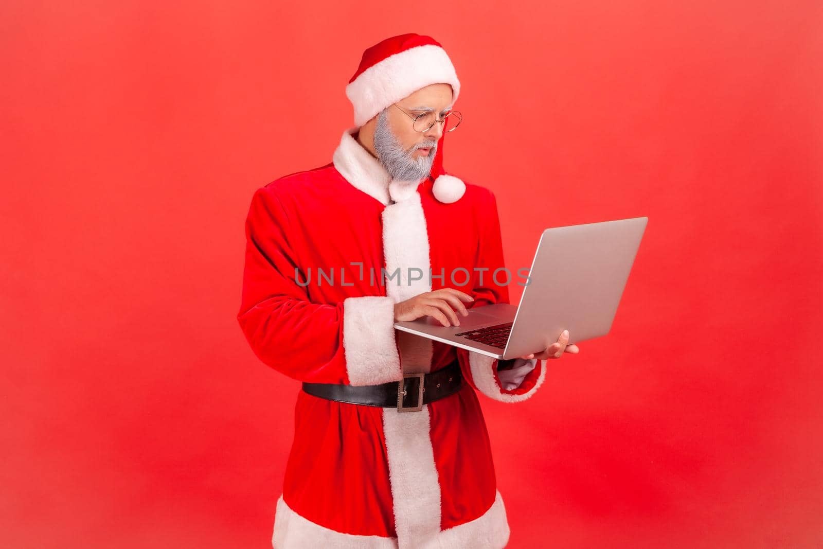 Serious elderly man with gray beard wearing santa claus costume working on computer, looking at display with concentrated facial expression. Indoor studio shot isolated on red background.