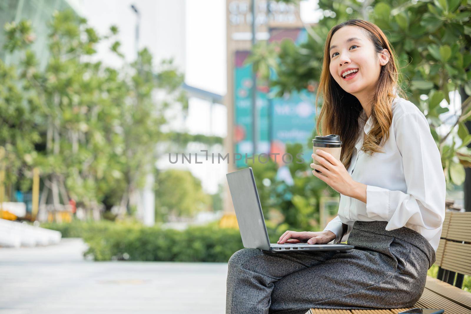 Happy Asian professional smiling businesswoman sitting alone typing computer outside street city by Sorapop