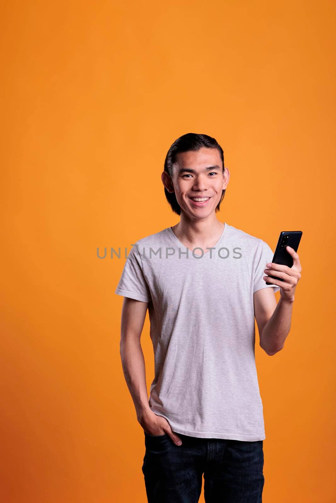 Young smiling asian man using smartphone, messaging on social media. Joyful teenager standing with happy facial expression, holding mobile phone, online communication concept